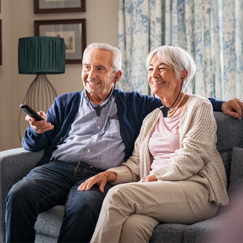 Senior couple sitting on a couch using wireless TV listening devices, smiling while enjoying clear and comfortable television sound together